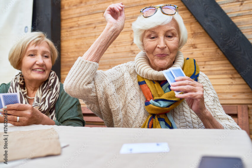 Portrait of cheerful seniors playing card game at lunch table on outdoor terrace, focus on two old women winning game
