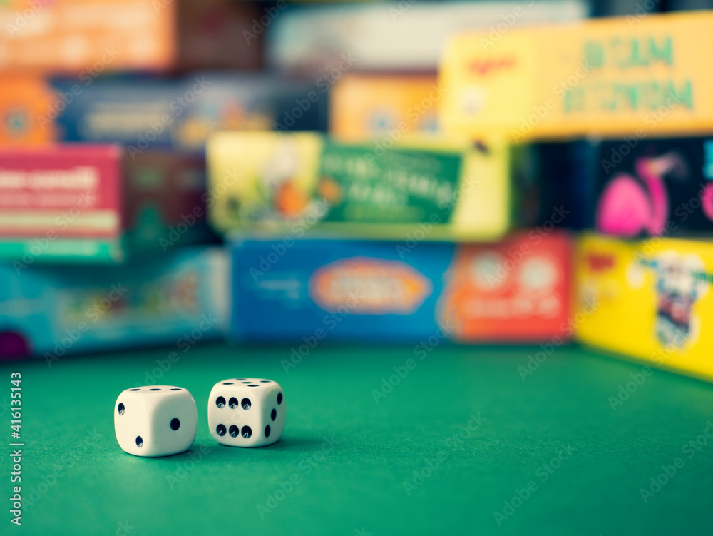 White dice on the green surface on the blurred background of colorful board game boxes. Concept of entertainment under lockdown